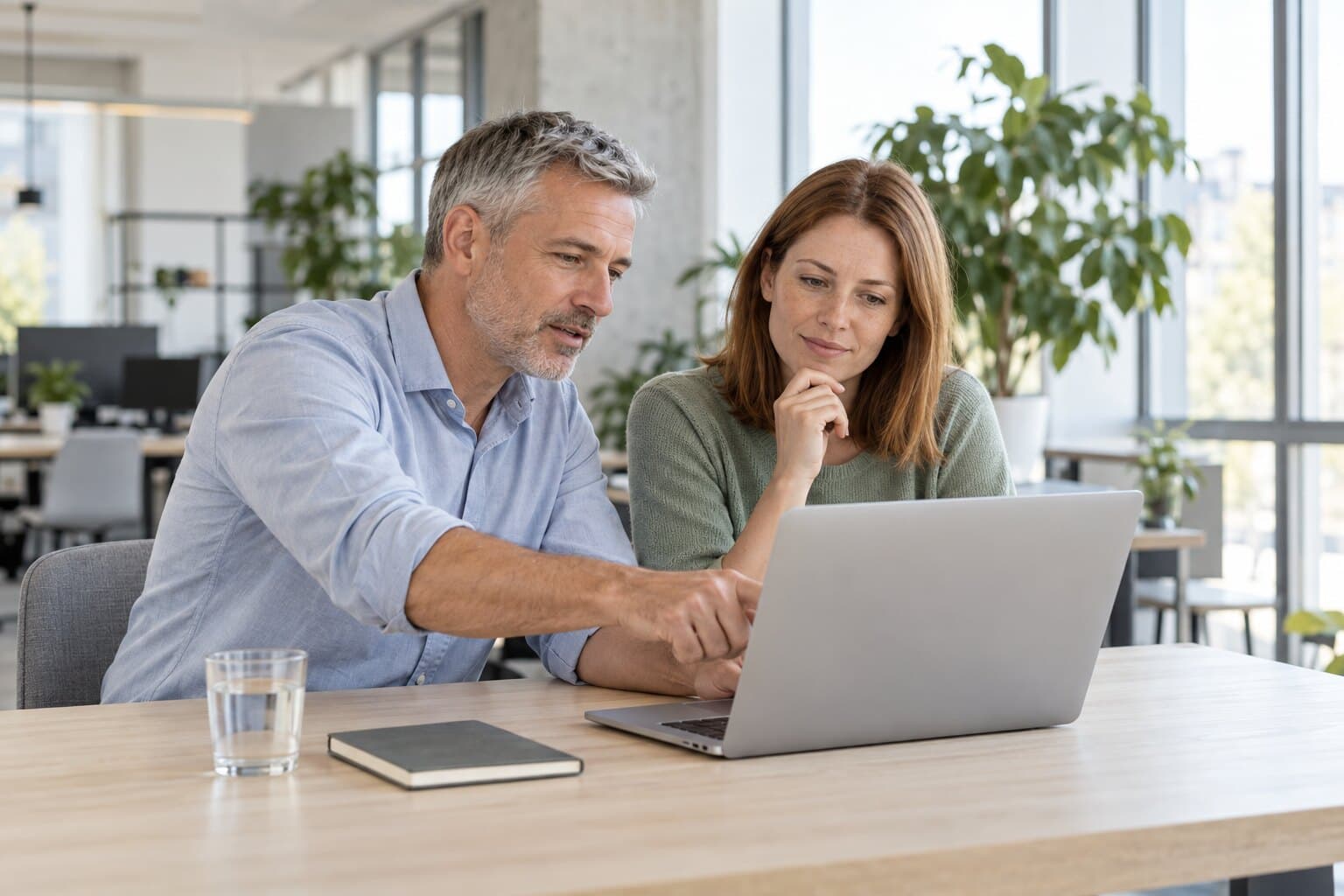 Two colleagues at a desk looking at one laptop, one pointing at the screen